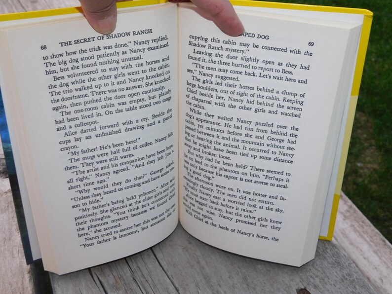 May include: Open book with yellow pages, showing a scene from a Nancy Drew mystery novel. The text reads "The Secret of Shadow Ranch" and "The men may come back. Let's wait here and see."