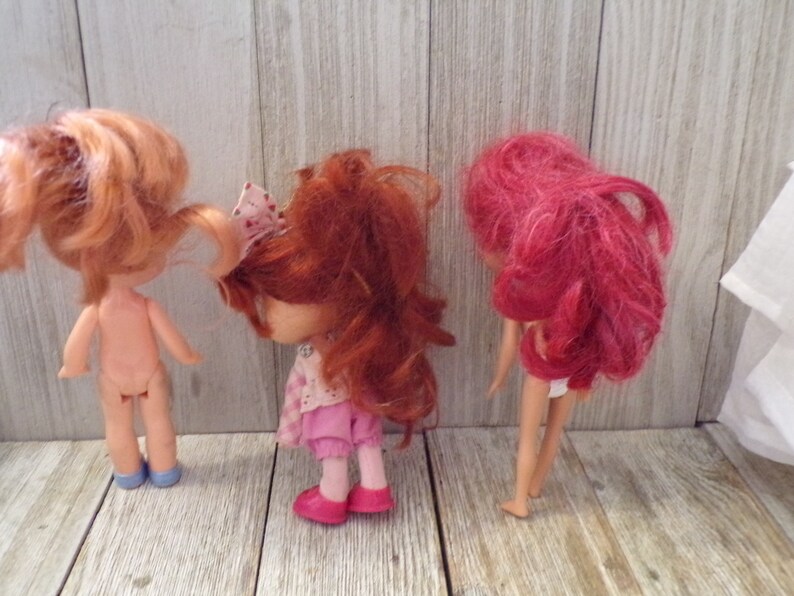 Pode incluir: Tr&ecirc;s bonecas com cabelo vermelho. A boneca &agrave; esquerda est&aacute; nua e usa sapatos azuis. A boneca do meio est&aacute; usando um vestido rosa e sapatos rosa. A boneca &agrave; direita est&aacute; usando um mai&ocirc; branco e tem cabelo rosa.