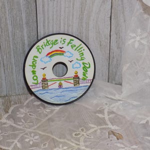 May include: A round, black disc with a hand-painted design of a bridge with a rainbow over it. The text "London Bridge is Falling Down" is written around the edge of the disc.