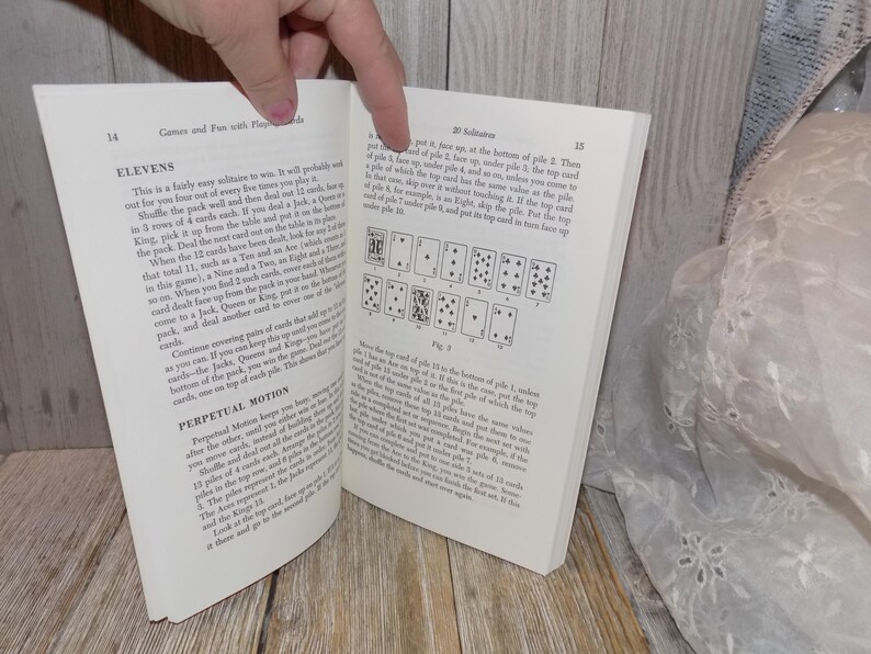 May include: A book titled "Games and Fun with Playing Cards" with an open page showing a card game called "Elevens" and a card game called "Solitaires". The page shows the rules for the games and a diagram of the card layout for the "Solitaires" game.