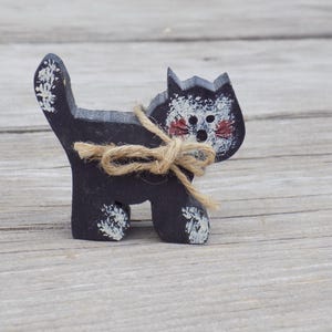 May include: A black wooden cat figurine with white painted details and a brown twine bow. The cat has a whimsical expression with red painted eyes.