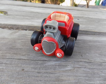 May include: A red toy car with a gray engine and a heart-shaped grill. The car has black wheels and a white interior.
