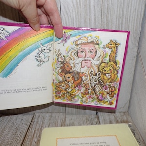 May include: An open children's book with a colorful illustration of a rainbow, a dove, and a man with a long beard surrounded by animals. The text on the page reads "From that day forth, all men who saw a rainbow knew of the promise of the Lord, and the great faith of a man called Noah."