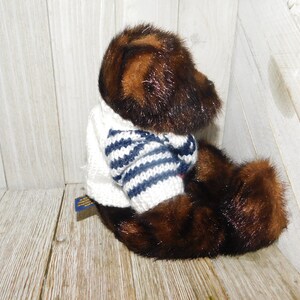 May include: A brown and black plush teddy bear wearing a white sweater with blue and white stripes. The bear is sitting on a wooden surface.