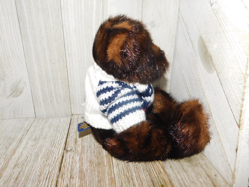 May include: A brown and black plush teddy bear wearing a white sweater with blue and white stripes. The bear is sitting on a wooden surface.