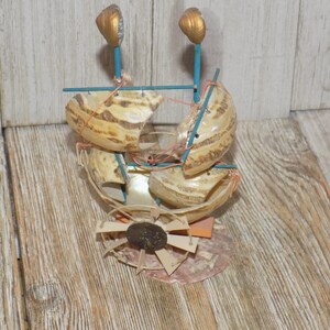 May include: A handcrafted seashell boat sculpture. The boat features beige shells, blue supports, and gold-painted accents. The design includes a fan-like structure at the base, with a dark brown stone center.