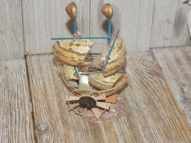 May include: A handcrafted seashell boat sculpture. The boat features beige shells, blue supports, and gold-painted accents. The design includes a fan-like structure at the base, with a dark brown stone center.