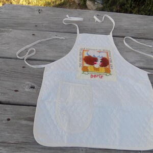 Op de afbeelding: Witte gewatteerde schort met een zak en een kleurrijk geborduurd patch met de tekst "This food from Doris Kitchen is sealed with love Doris".
