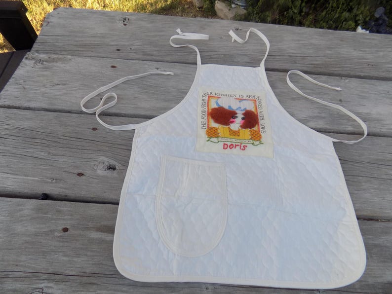 Op de afbeelding: Witte gewatteerde schort met een zak en een kleurrijk geborduurd patch met de tekst "This food from Doris Kitchen is sealed with love Doris".