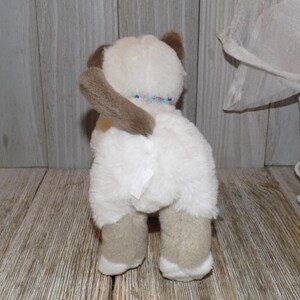 May include: A plush toy cat with white fur, brown ears, and tan paws. The cat has a brown tail and a colorful beaded collar. The toy is positioned on a wooden surface.
