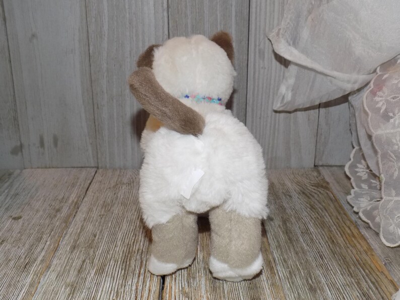 May include: A plush toy cat with white fur, brown ears, and tan paws. The cat has a brown tail and a colorful beaded collar. The toy is positioned on a wooden surface.
