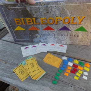 May include: A vintage board game called "Bibleopoly" with a purple, blue, and green background. The game board is made of cardboard and has a stone-like texture. The game pieces are colorful plastic houses and a set of yellow tickets.