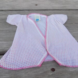 May include: A white baby gown with pink polka dots and pink trim. The gown has a snap closure at the neckline and is made of a soft, comfortable fabric.