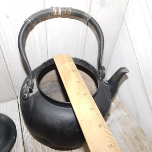May include: Black cast iron kettle with a black woven handle and a wooden ruler for scale. The kettle has a spout and a lid.