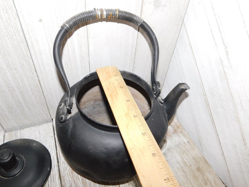 May include: Black cast iron kettle with a black woven handle and a wooden ruler for scale. The kettle has a spout and a lid.