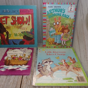May include: Four children's books with colorful covers. The first book is blue and features a black and brown illustration of a pet show. The second book is red, yellow, and green and features a cartoon illustration of Arthur from the Arthur book series. The third book is purple and features a colorful illustration of Noah's Ark. The fourth book is yellow and features a cartoon illustration of a raccoon.