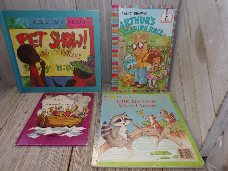 May include: Four children's books with colorful covers. The first book is blue and features a black and brown illustration of a pet show. The second book is red, yellow, and green and features a cartoon illustration of Arthur from the Arthur book series. The third book is purple and features a colorful illustration of Noah's Ark. The fourth book is yellow and features a cartoon illustration of a raccoon.