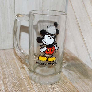 May include: A clear glass Mickey Mouse mug with a black outline and red shorts. The mug has a handle and says "Mickey Mouse" and "Walt Disney Productions" on the bottom.
