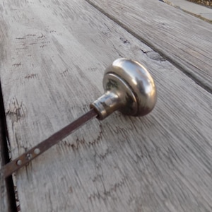 May include: An antique, silver-toned door knob with a rounded handle and a rectangular, rusty stem. The knob is resting on a weathered, gray wooden surface, showcasing its vintage aesthetic.