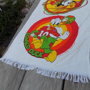 Peut inclure: Serviette en coton blanc avec un motif color&eacute; de Donald Duck en uniforme de football. La serviette a un bord &agrave; franges et un design r&eacute;tro avec les mots "Donald Duck" en cercle.