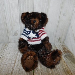 May include: A brown teddy bear wearing a red, white and blue knitted sweater with a star design. The bear is sitting on a wooden surface.
