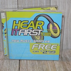 May include: A CD case with a blue and green design. The text "HEAR IT FIRST.com" is displayed with a graphic of headphones. The words "NEW MUSIC SAMPLER" and "FREE OR $2.99" are also visible.
