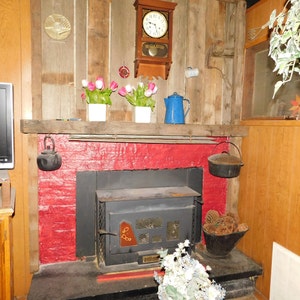 May include: A red brick fireplace with a black wood-burning stove. The stove has a glass door and a metal grate. There is a wooden mantel above the fireplace with a clock, flowers, and a blue enamel teapot.