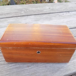 May include: A rectangular wooden box with a hinged lid. The box is made of reddish-brown wood with a glossy finish. A small, round metal clasp is centered on the front. The box is sitting on a weathered wooden surface.