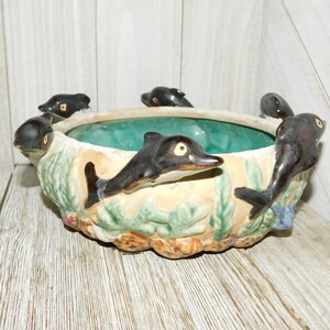 May include: A ceramic planter shaped like a bowl with a green glazed interior. The exterior is decorated with a scene of dolphins swimming in the ocean, with seaweed and seashells.