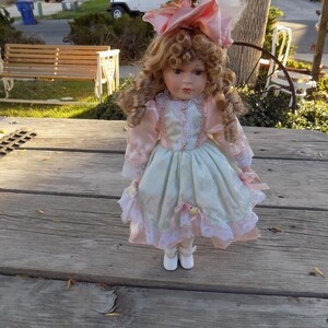 May include: A porcelain doll with blonde curly hair, wearing a pink and light blue dress with lace trim and a matching bow. The doll is standing on a weathered wooden surface.