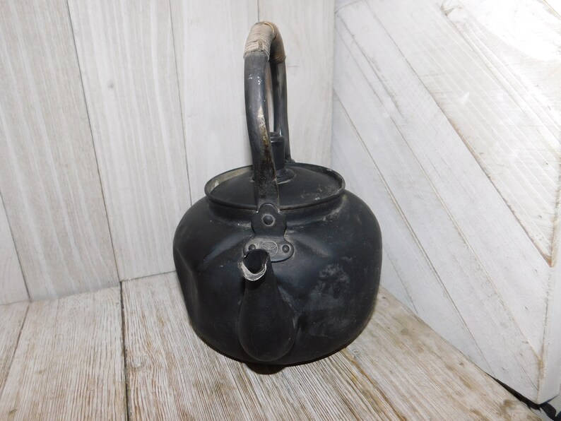 May include: A black metal tea kettle with a wooden handle. The kettle has a dented and worn appearance.