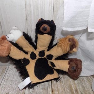 May include: A brown and black plush hand puppet with five different animal heads. The animal heads include a deer, a bear, a dog, a cat, and a rabbit. The puppet has a large paw print on the palm.