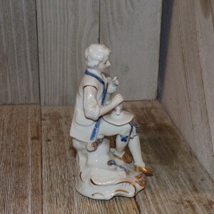 Pu&ograve; includere: Una statuetta in porcellana di un uomo in un abito blu e bianco seduto su uno scoglio. La statuetta &egrave; dipinta con accenti dorati e ha un design dettagliato.