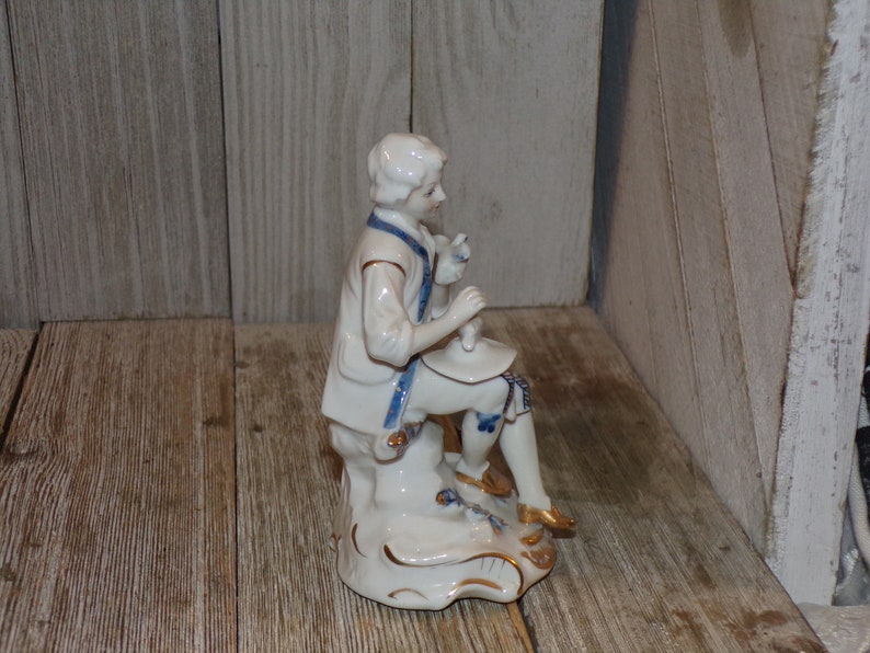 Pu&ograve; includere: Una statuetta in porcellana di un uomo in un abito blu e bianco seduto su uno scoglio. La statuetta &egrave; dipinta con accenti dorati e ha un design dettagliato.