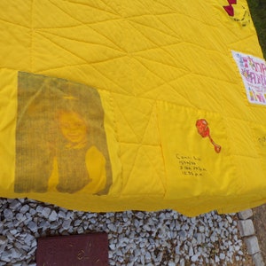 May include: A handmade yellow quilt with a diamond pattern. It features a printed image of a person, a rattle illustration, and handwritten text with a name and birth details. Other patches include text and a basket illustration.