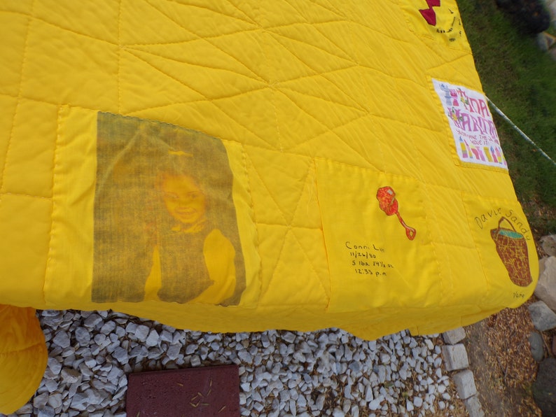 May include: A handmade yellow quilt with a diamond pattern. It features a printed image of a person, a rattle illustration, and handwritten text with a name and birth details. Other patches include text and a basket illustration.