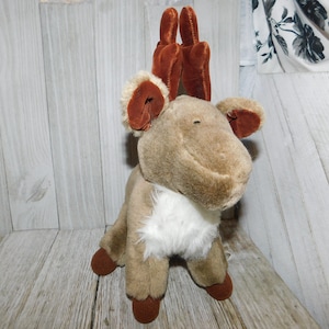 May include: A plush brown reindeer toy with white fur on its chest and brown velvet antlers. The reindeer is sitting on a wooden surface.