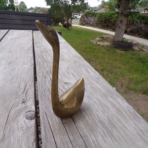 Pu&ograve; includere: Una statuetta di cigno in ottone dorato con un collo lungo e un corpo curvo. Il cigno &egrave; seduto su una superficie di legno.