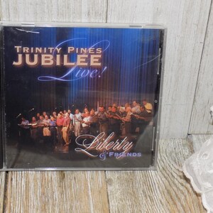May include: A CD case featuring the album "Trinity Pines Jubilee Live!" with the text "Liberty & Friends." The cover shows a group of people on a stage with a blue backdrop. The CD case is in a clear plastic case.