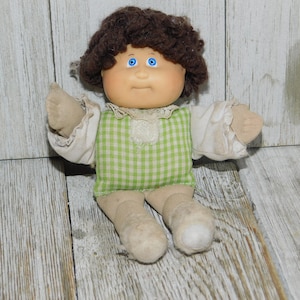 May include: A vintage doll with a light skin tone, blue eyes, and brown curly hair. The doll wears a green and white checkered dress with white sleeves and a lace collar. The doll's arms and legs are made of a soft, beige fabric.