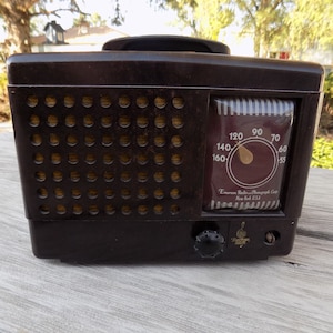 Puede incluir: Una radio Emerson marr&oacute;n vintage con una rejilla de altavoz y un dial de sintonizaci&oacute;n. El dial muestra n&uacute;meros del 55 al 160. La radio tiene un asa en la parte superior y un bot&oacute;n de sintonizaci&oacute;n en la parte frontal. El texto "Emerson Radio-Phonograph Corp New York USA" es visible.