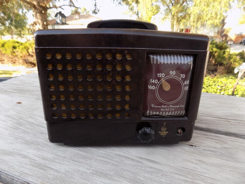 Puede incluir: Una radio Emerson marr&oacute;n vintage con una rejilla de altavoz y un dial de sintonizaci&oacute;n. El dial muestra n&uacute;meros del 55 al 160. La radio tiene un asa en la parte superior y un bot&oacute;n de sintonizaci&oacute;n en la parte frontal. El texto "Emerson Radio-Phonograph Corp New York USA" es visible.