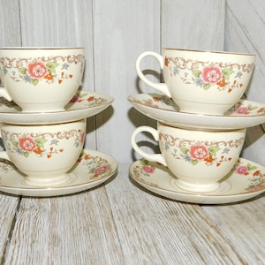 May include: Four cream-colored teacups and saucers. Each cup and saucer features a floral design with pink roses, orange accents, and gold trim. The cups are stacked in pairs, creating a classic tea set.