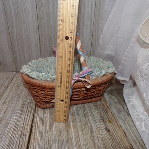 Peut inclure: Un petit panier en osier avec une doublure florale bleu et blanc. Le panier a une poign&eacute;e envelopp&eacute;e d'un ruban bleu et rose. Le panier est pos&eacute; sur une surface en bois.