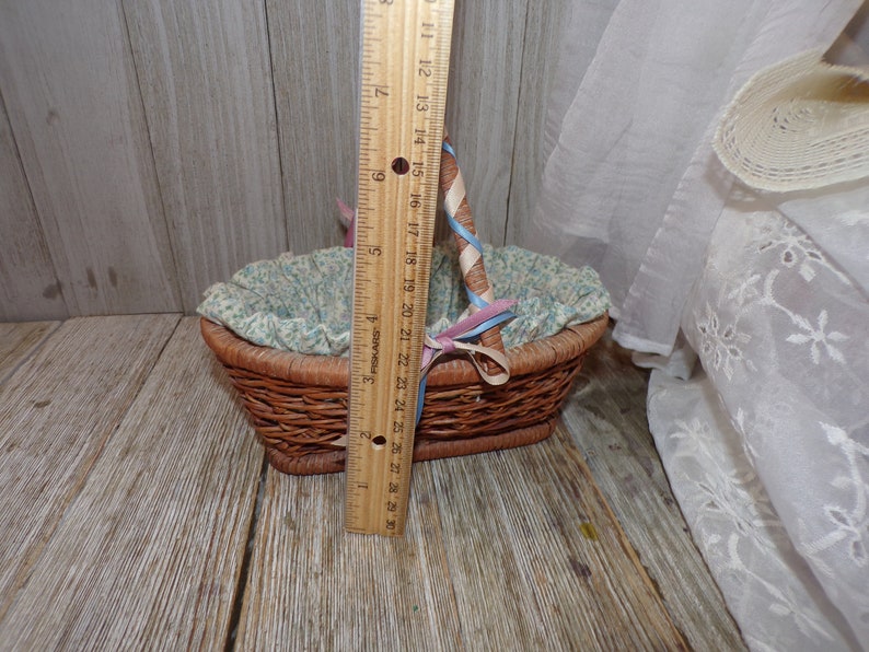 Peut inclure: Un petit panier en osier avec une doublure florale bleu et blanc. Le panier a une poign&eacute;e envelopp&eacute;e d'un ruban bleu et rose. Le panier est pos&eacute; sur une surface en bois.