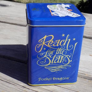 May include: Blue metal tin with gold lettering that reads "Reach for the Stars!" and "Pocket Dragons".