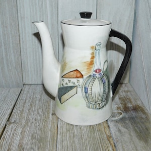 May include: A white ceramic coffee pot with a black handle and lid. The pot has a painted design of a bottle of wine, a wedge of cheese, and a block of cheese.