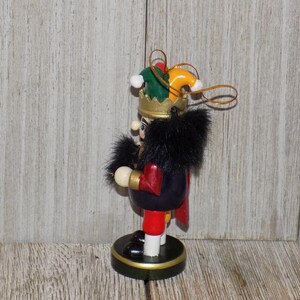May include: A wooden nutcracker ornament with a black fur hat and a green and yellow jester hat. The nutcracker is wearing a red and black suit and holding a gold scepter. The ornament is hanging from a gold string.