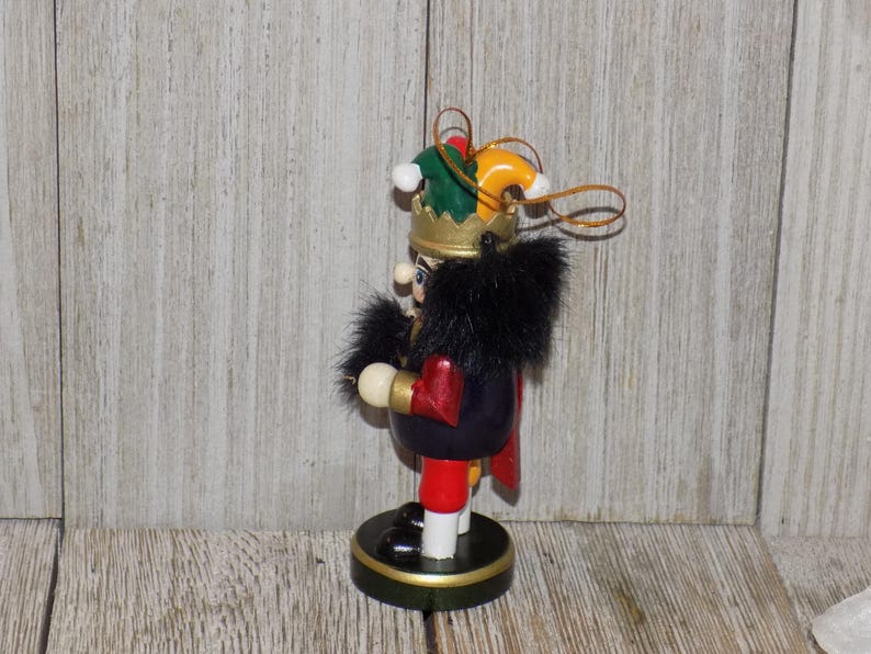 May include: A wooden nutcracker ornament with a black fur hat and a green and yellow jester hat. The nutcracker is wearing a red and black suit and holding a gold scepter. The ornament is hanging from a gold string.
