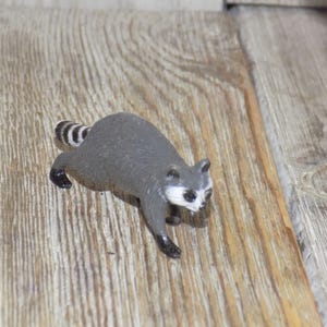 May include: A gray and white raccoon figurine with a black tail and a black nose. The figurine is sitting on a wooden surface.